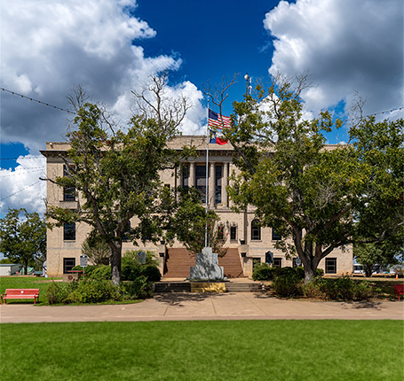 Historic government building exterior with American and Texas flags surrounded by large trees and landscaped grounds